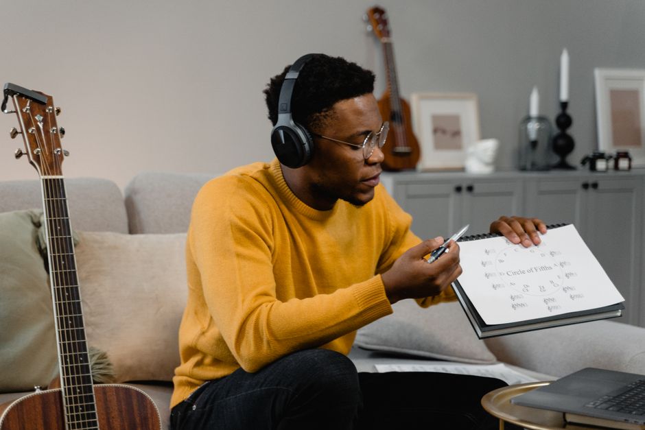 A man wearing headphones studies music theory on a sofa with a guitar, laptop, and notebook.